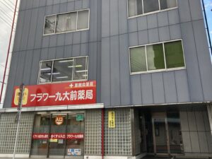 福岡事務所の外観。ヤマモトビルディングはブルーグレーの外観をしており、小さな入り口を備えています。ビル1階には「フラワー九大前薬局」があります。
