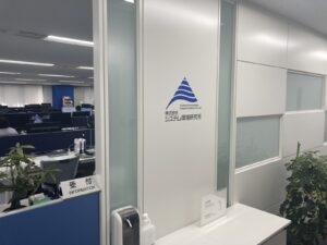 東京事務所の入口。エレベータで２階に上がり、正面の扉を開けると、当社ロゴをあしらった間仕切り壁と受付があります。